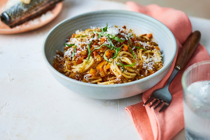 A white bowl of spaghetti bolognese with parmesan and a grater in the background