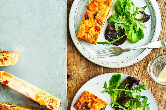 A slice of frittata with green leaves on a grey plate, set against a wooden board