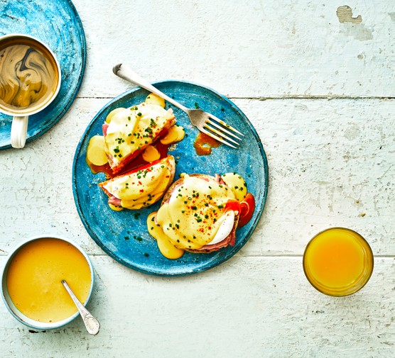 A white-washed background topped with blue plates, with two English muffins, poached eggs and hollandaise sauce on top. There is a cup of coffee and glass of orange juice on the table