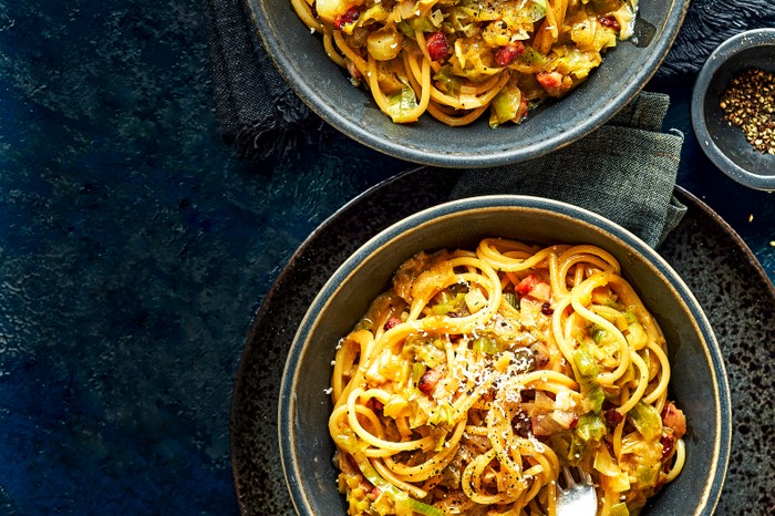 Two grey bowls filled with spaghetti, fried leeks and chunks of fried pancetta, topped with grated white cheese