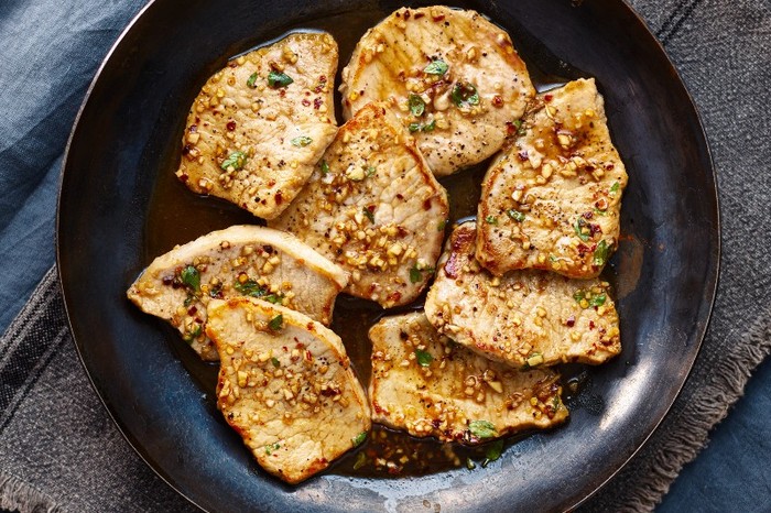 A frying pan with pork medallions in a glossy, light sauce on a grey linen napkin