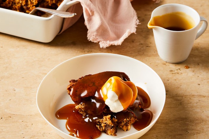 A bowl of sticky pudding topped with a dollop of cream, plus a jug of toffee sauce and a baking tray of sticky toffee pudding