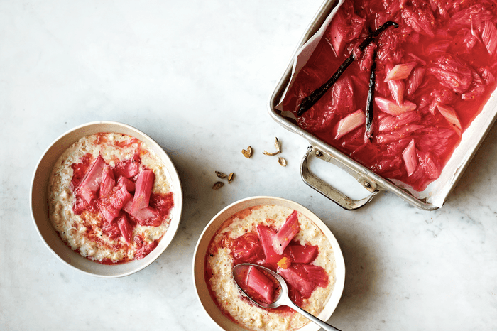 A tray of baked rhubarb with vanilla pods in an oven tray, next to two bowls of rhubarb-topped porridge