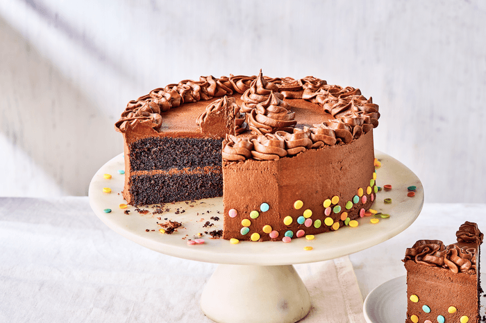 A chocolate frosted cake on a white cake stand with a slice cut out of it in front of a white background